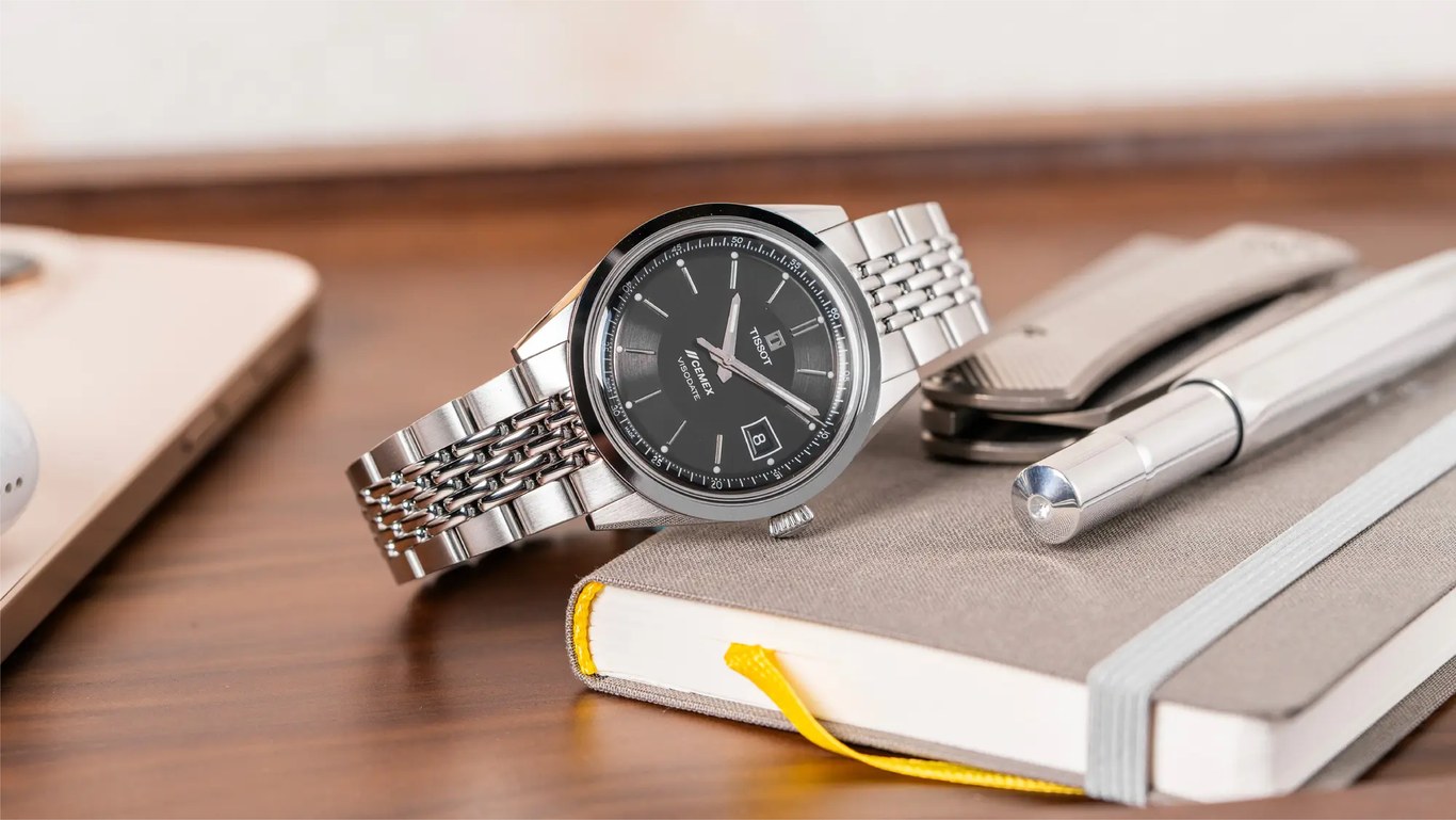 Reloj Tissot Visodate de acero con carátula negra y el logotipo de CEMEX, apoyado sobre una libreta gris junto a una pluma, en una mesa de madera.